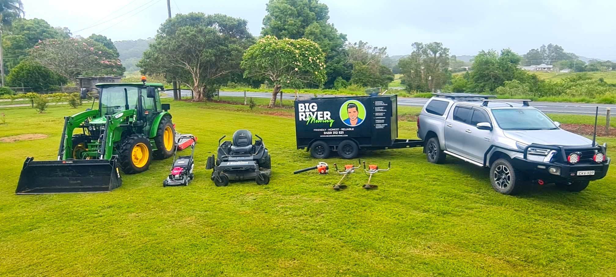 Big River Mowing's full kit on a Lower Clarence lawn — green John Deere tractor with loader, two ride-on mowers, push mower, Stihl whippers and blowers laid out beside the silver Hilux and the branded trailer.