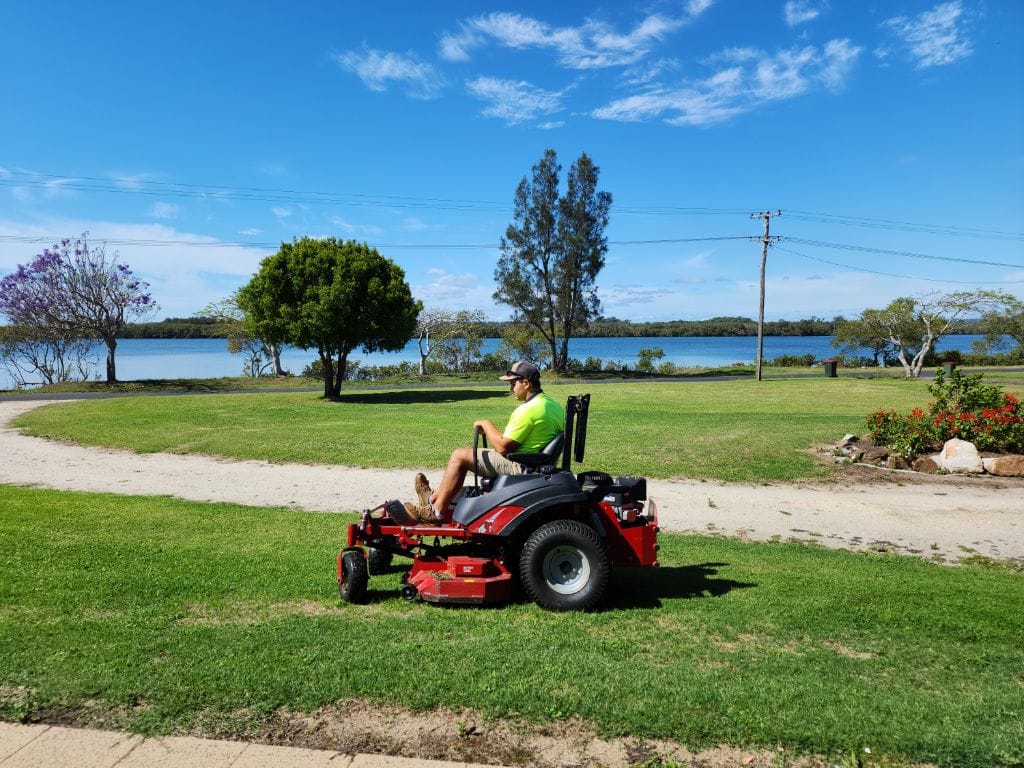 Steven on the red zero-turn mower beside the Clarence River — jacaranda in flower, calm water, neatly cut bank.